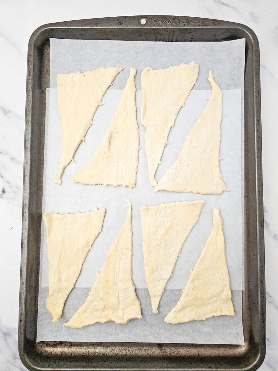 A sheet of parchment paper laid on a dark baking pan holds eight triangles of unrolled crescent dough, arranged neatly in rows.
