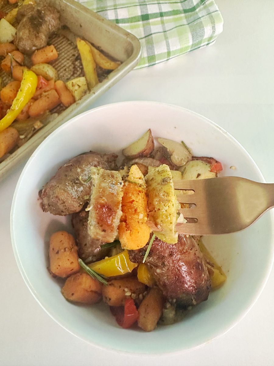  A fork holding a bite of roasted sausage, carrot, and potato, with a serving bowl of the finished meal in the background.