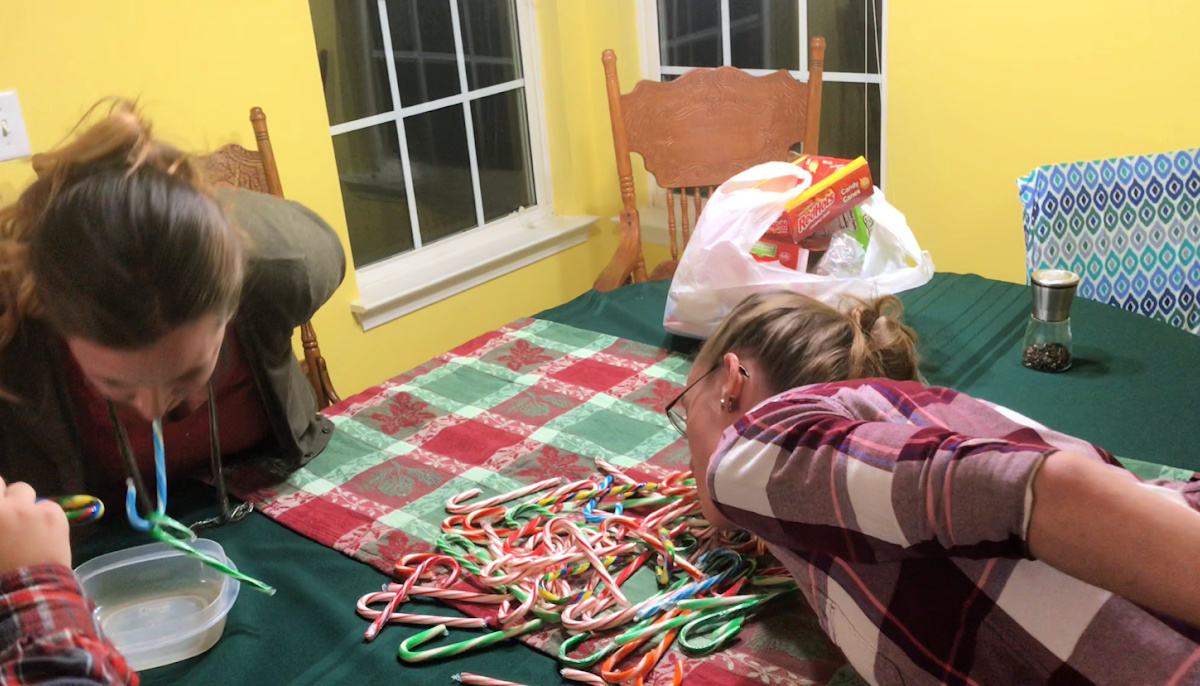 Two players, leaning over a green and red tartan tablecloth, with their faces close to a pile of candy canes, using the hook of a cane held in their mouth to try and pick up or "bob" another cane for the Christmas party game.