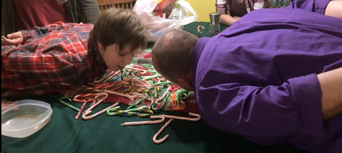 A close-up action shot of two players, leaning over a table covered in colorful candy canes, actively using a cane held in their mouths to "fish" or hook other candy canes