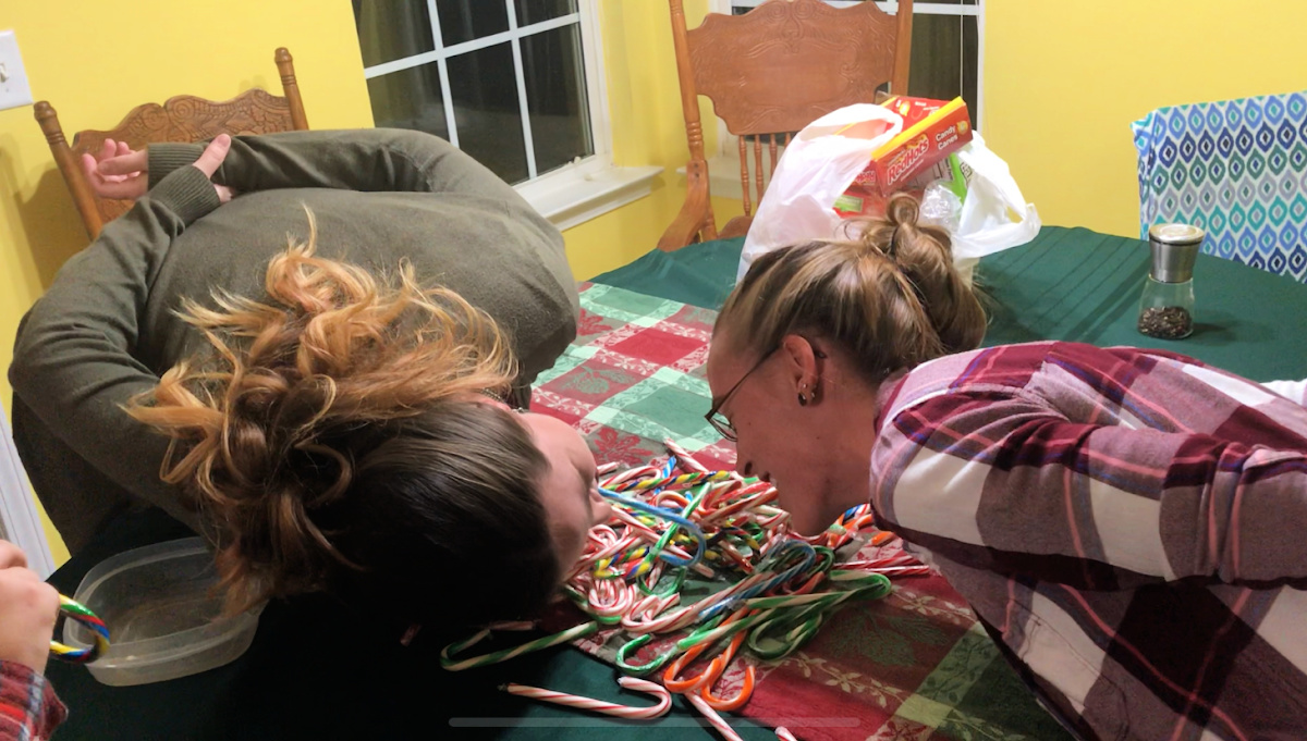 A hilarious action photo of two young adults competing in the Bobbing for Candy Canes Christmas party game, leaning over a table full of multicolored candy canes and laughing while attempting to hook the candy with a cane in their mouth.