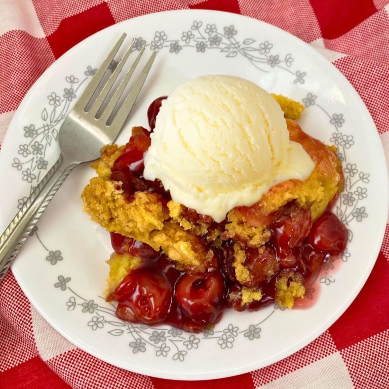 A slice of warm cherry dump cake, topped with a scoop of melting vanilla ice cream, sits on a white plate with a floral pattern. A silver fork rests on the left side of the plate, which is set against a red and white checkered tablecloth. The dessert features bubbly, bright red cherries and a golden-brown, slightly crumbly cake topping.