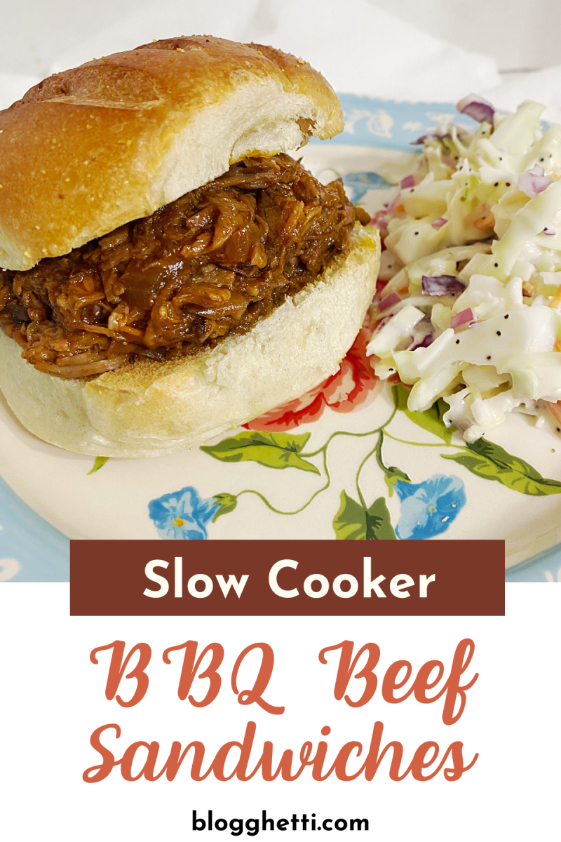 A plate with a slow cooker BBQ beef sandwich on a bun and a side of coleslaw. The sandwich has a generous amount of shredded beef covered in a thick, dark sauce. The coleslaw is ion a blue plate and is made with shredded cabbage and carrots.