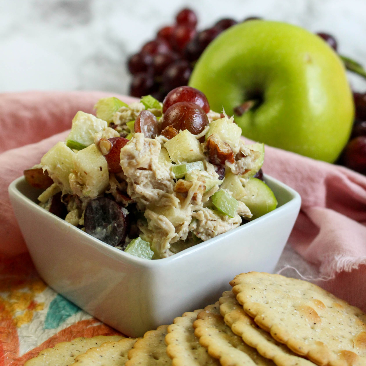 close up of a bowl with fancy chicken salad with fruit. Surronded by an apple and grapes with crackers