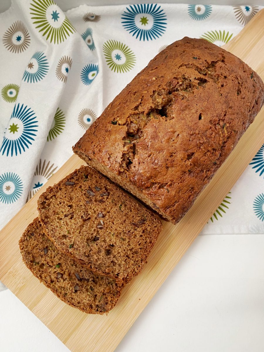 An overhead view of a whole zucchini nut bread loaf and two slices on a light wooden cutting board.
