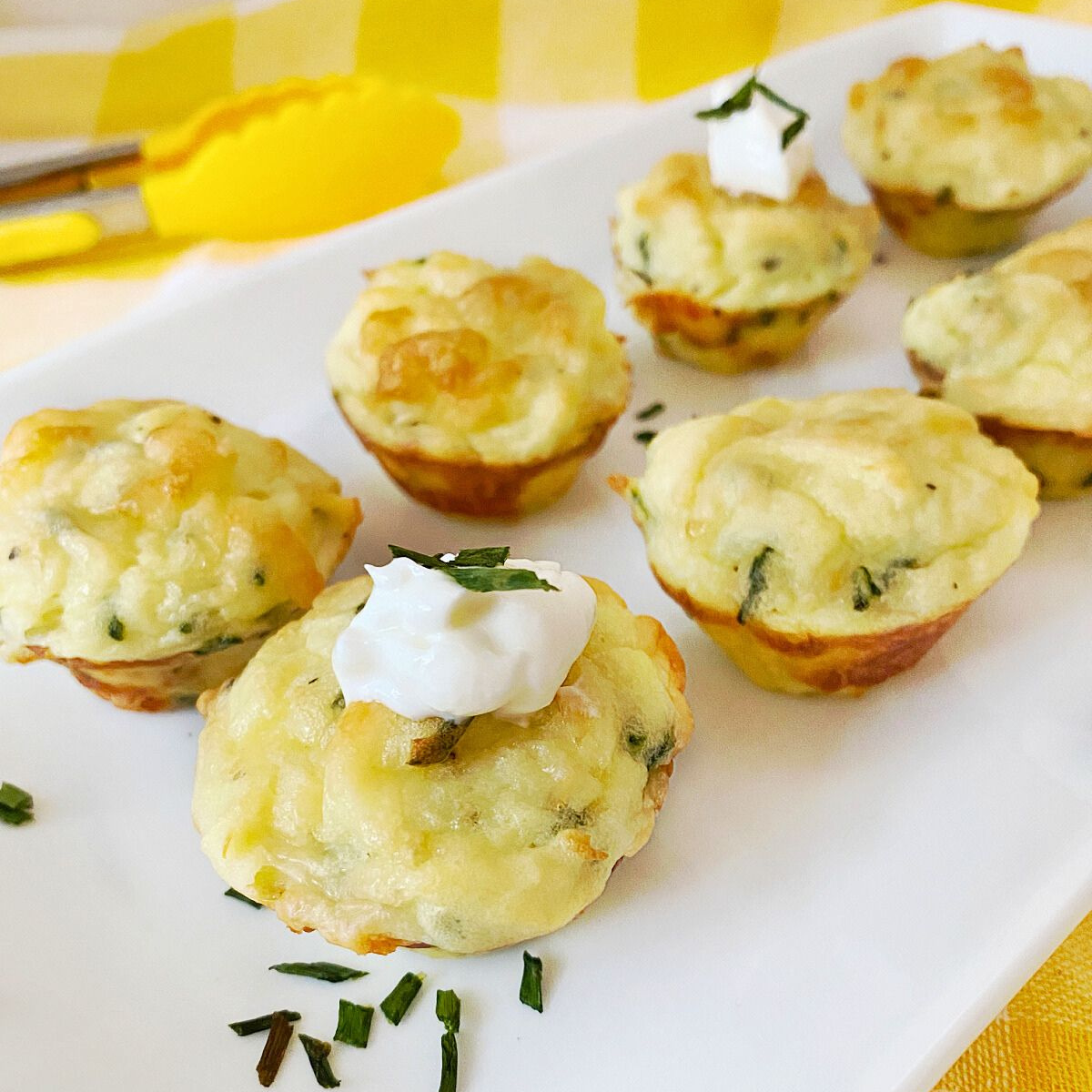 A group of golden-brown mashed potato puffs are arranged on a white rectangular plate. One in the foreground is topped with sour cream and a sprig of chives. Yellow tongs and a yellow and white checked napkin are visible in the background.