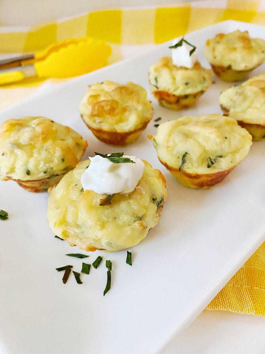 A white serving tray holds several baked mashed potato puffs, some garnished with sour cream and chives, sitting on a yellow and white checked cloth. Yellow tongs are visible in the background.