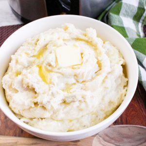 A square image shows a white bowl of creamy mashed potatoes with a melting pat of butter on top, set against a wooden board. A slow cooker and a green and white plaid napkin are visible in the background.