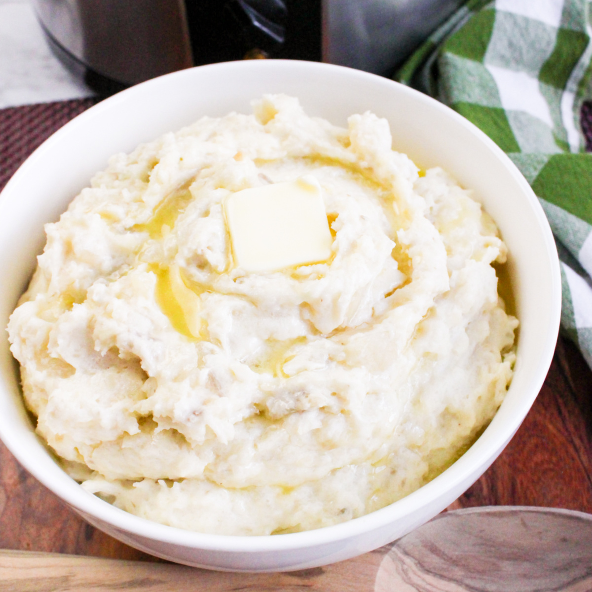 A square image shows a white bowl of creamy mashed potatoes with a melting pat of butter on top, set against a wooden board. A slow cooker and a green and white plaid napkin are visible in the background.