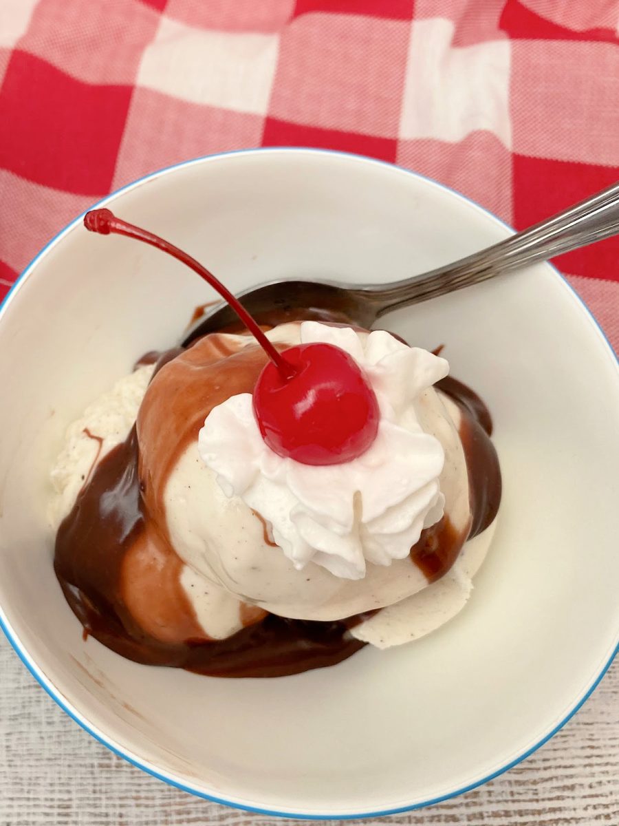 A scoop of vanilla bean ice cream topped with rich chocolate drizzle, whipped cream, and a bright red maraschino cherry.