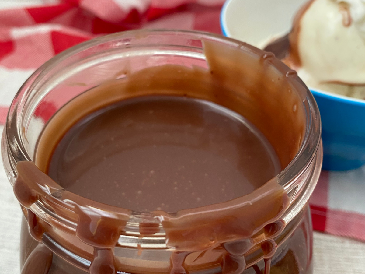 Close up view of a glass jar overflowing with warm, creamy chocolate ganache topping.