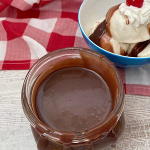 A glass jar filled with pourable chocolate fudge sitting on a white wooden surface with a sundae in the background.