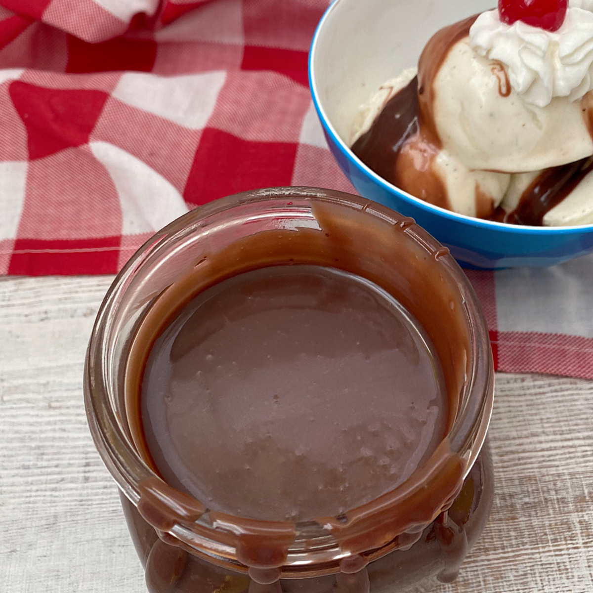 A glass jar filled with pourable chocolate fudge sitting on a white wooden surface with a sundae in the background.