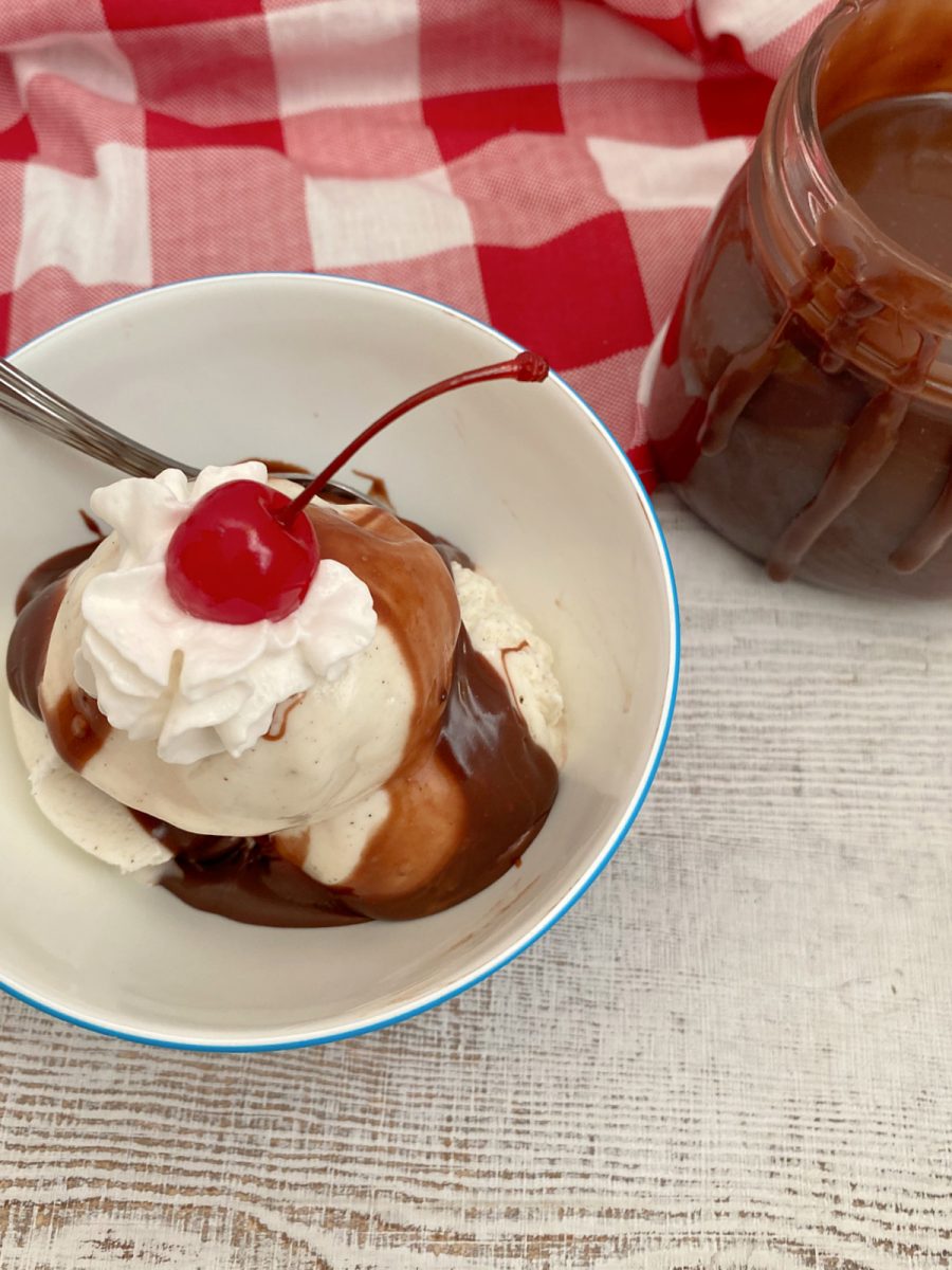 A bowl of vanilla bean ice cream drizzled with hot fudge and topped with whipped cream and a cherry, sitting next to a jar of extra sauce.