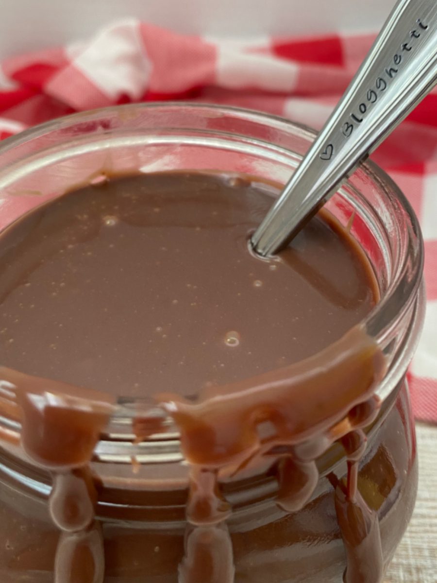 A metal spoon engraved with "Blogghetti" dipping into a glass jar filled with warm, glossy chocolate topping.