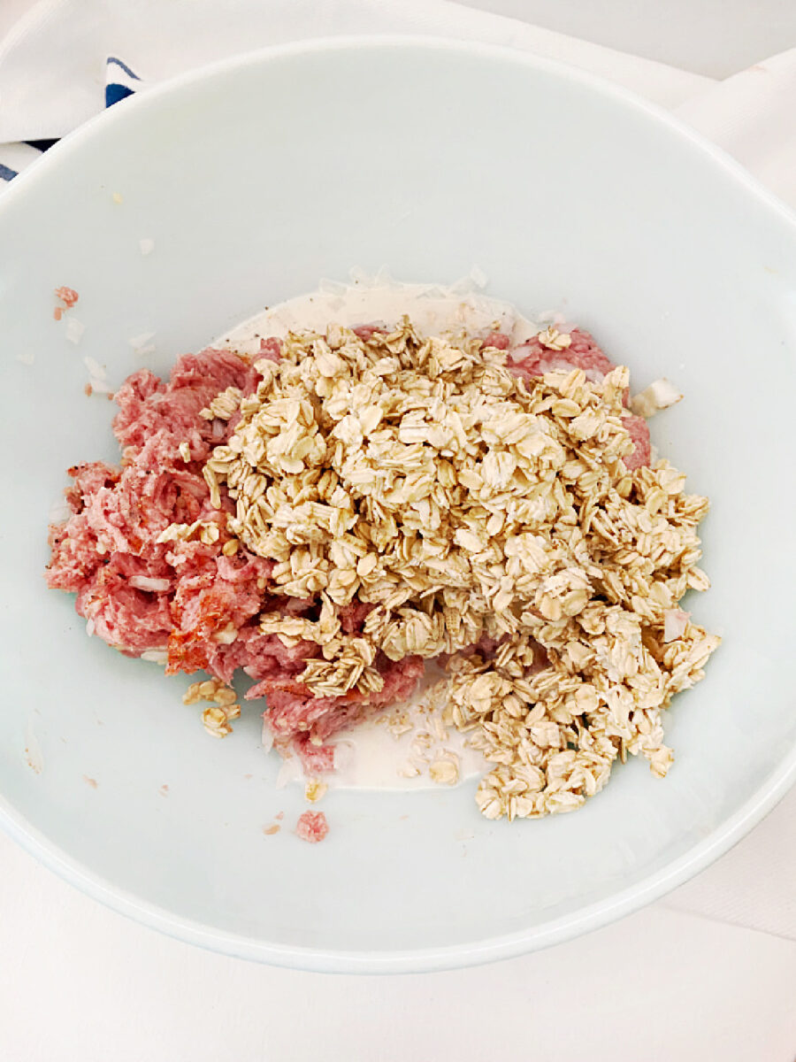 Rolled oats being added to seasoned ground turkey in a white mixing bowl for Mini Turkey Meatloaves.
