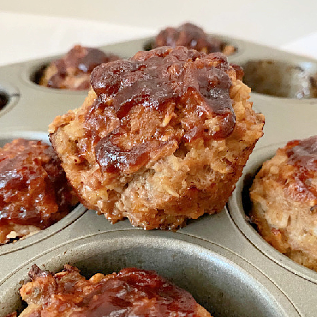 Close up of a BBQ glazed Mini Turkey Meatloaf lifted from a muffin tin with additional meatloaves in the background.
