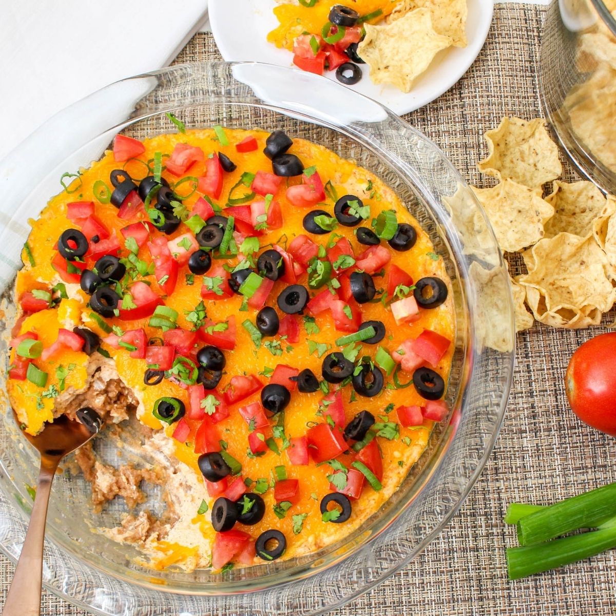 Hot taco dip baked in a glass pie dish with melted cheddar cheese, fresh toppings, and a serving spoon ready for scooping.