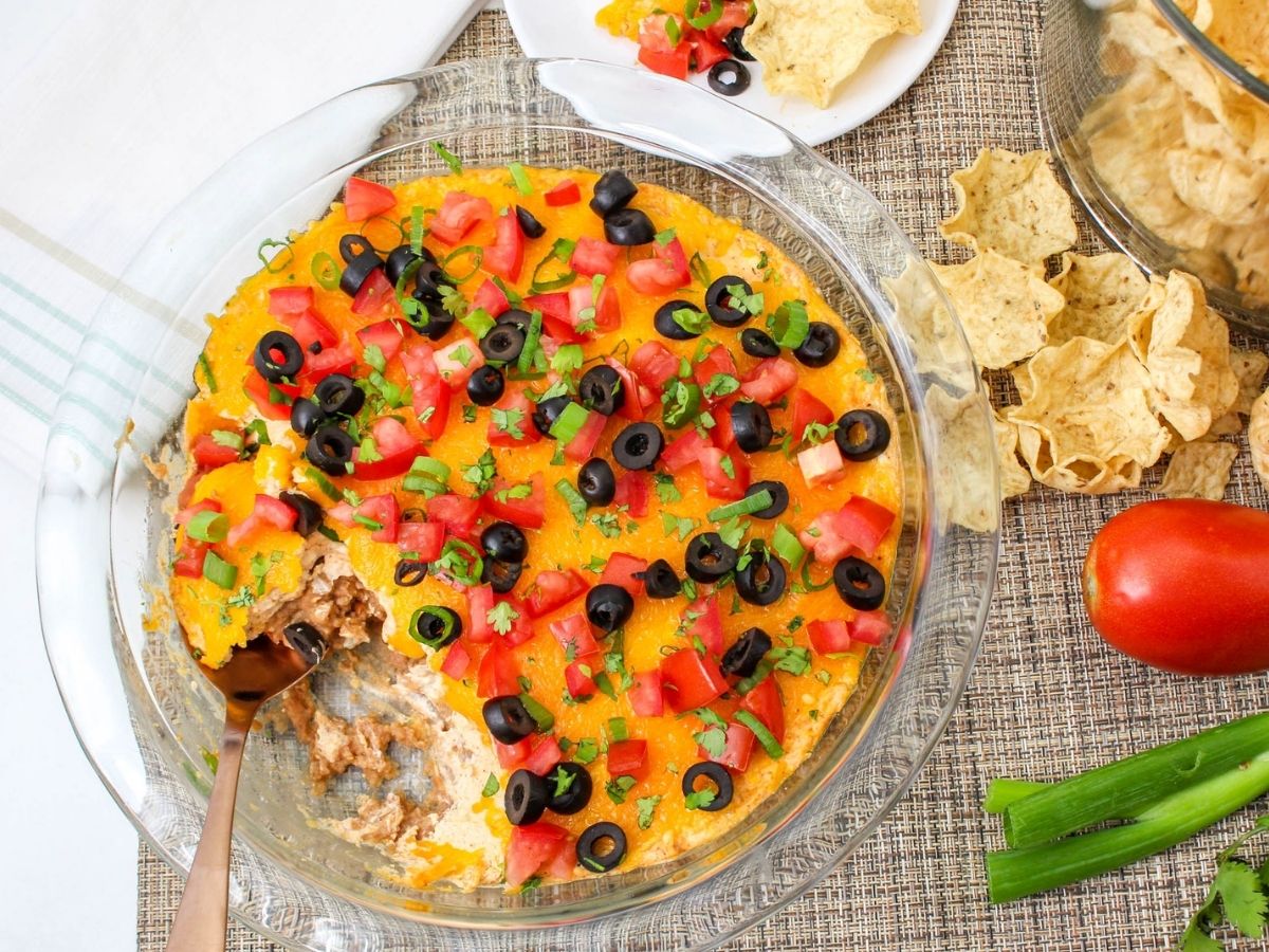 Overhead view of the baked dip topped with tomatoes, black olives, green onions, and cilantro with tortilla chips alongside.