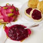 A vibrant whole pink dragon fruit sits next to a dragon fruit skin bowl filled with deep magenta pitaya jam. In the background, two golden-brown biscuits are topped with a generous spread of the seed-flecked fruit preserves on a white plate.
