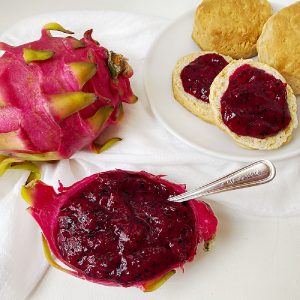 A vibrant whole pink dragon fruit sits next to a dragon fruit skin bowl filled with deep magenta pitaya jam. In the background, two golden-brown biscuits are topped with a generous spread of the seed-flecked fruit preserves on a white plate.