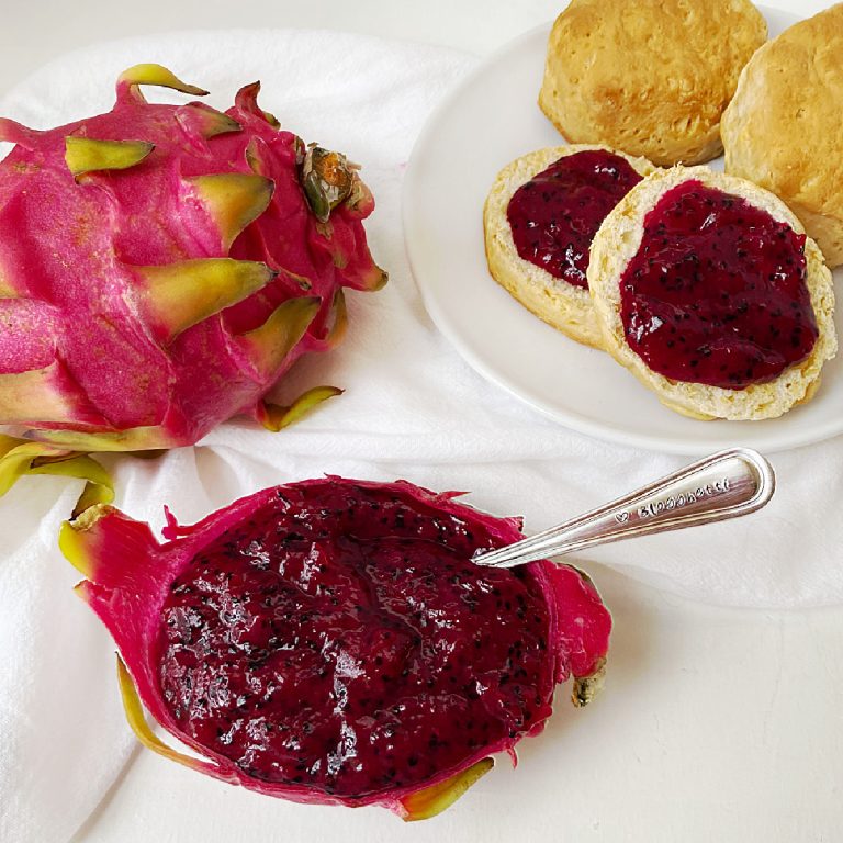 A vibrant whole pink dragon fruit sits next to a dragon fruit skin bowl filled with deep magenta pitaya jam. In the background, two golden-brown biscuits are topped with a generous spread of the seed-flecked fruit preserves on a white plate.