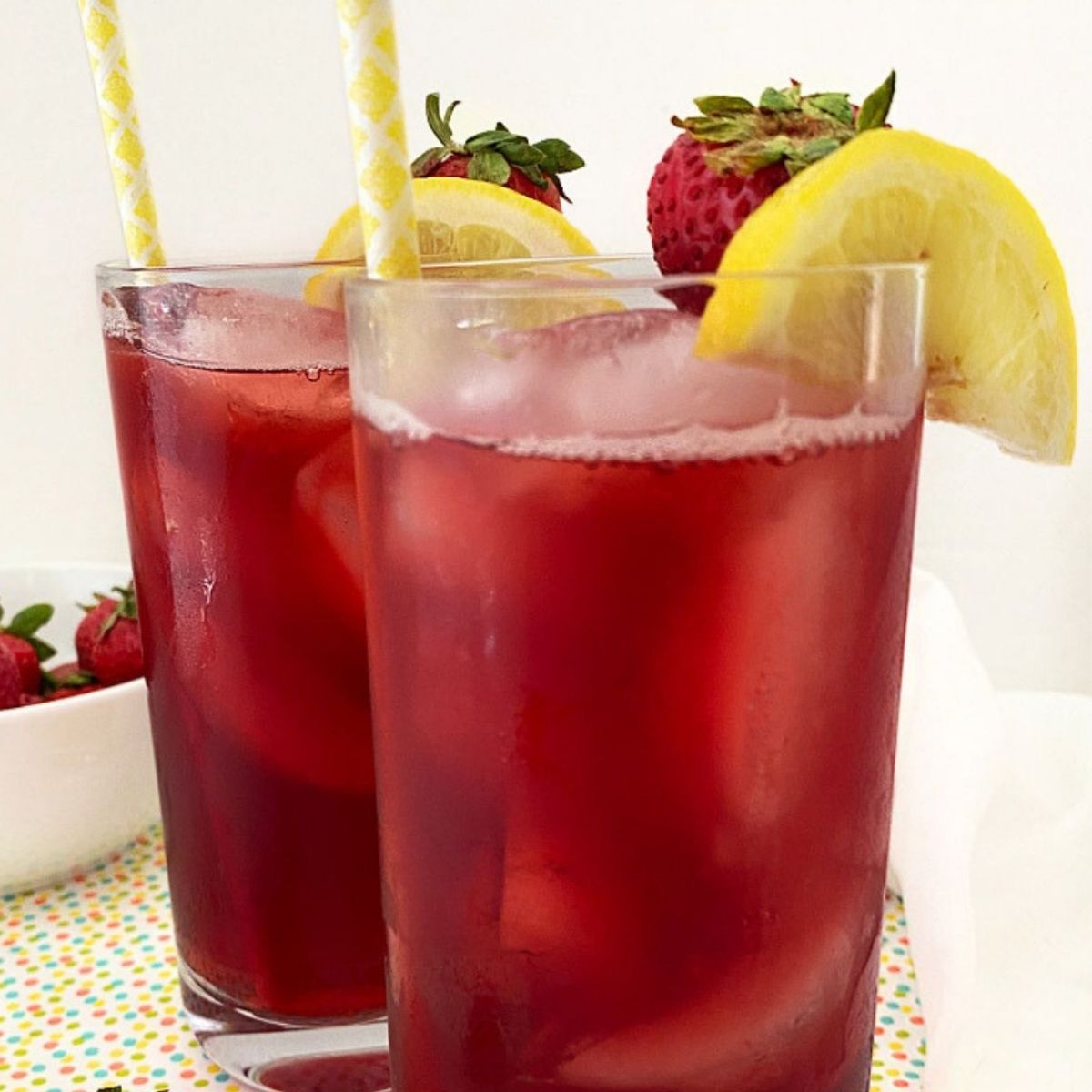 Two glasses of Strawberry Hibiscus Iced Tea filled with ice and garnished with fresh strawberries and lemon slices.