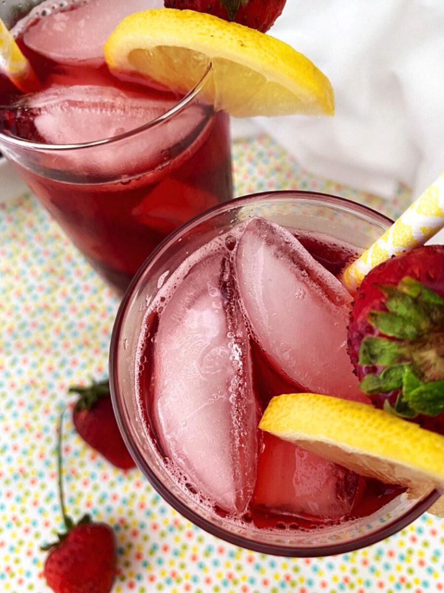 Overhead view of two glasses of Strawberry Hibiscus Iced Tea with ice, fresh strawberry garnish, lemon slices, and paper straws on a colorful tabletop.