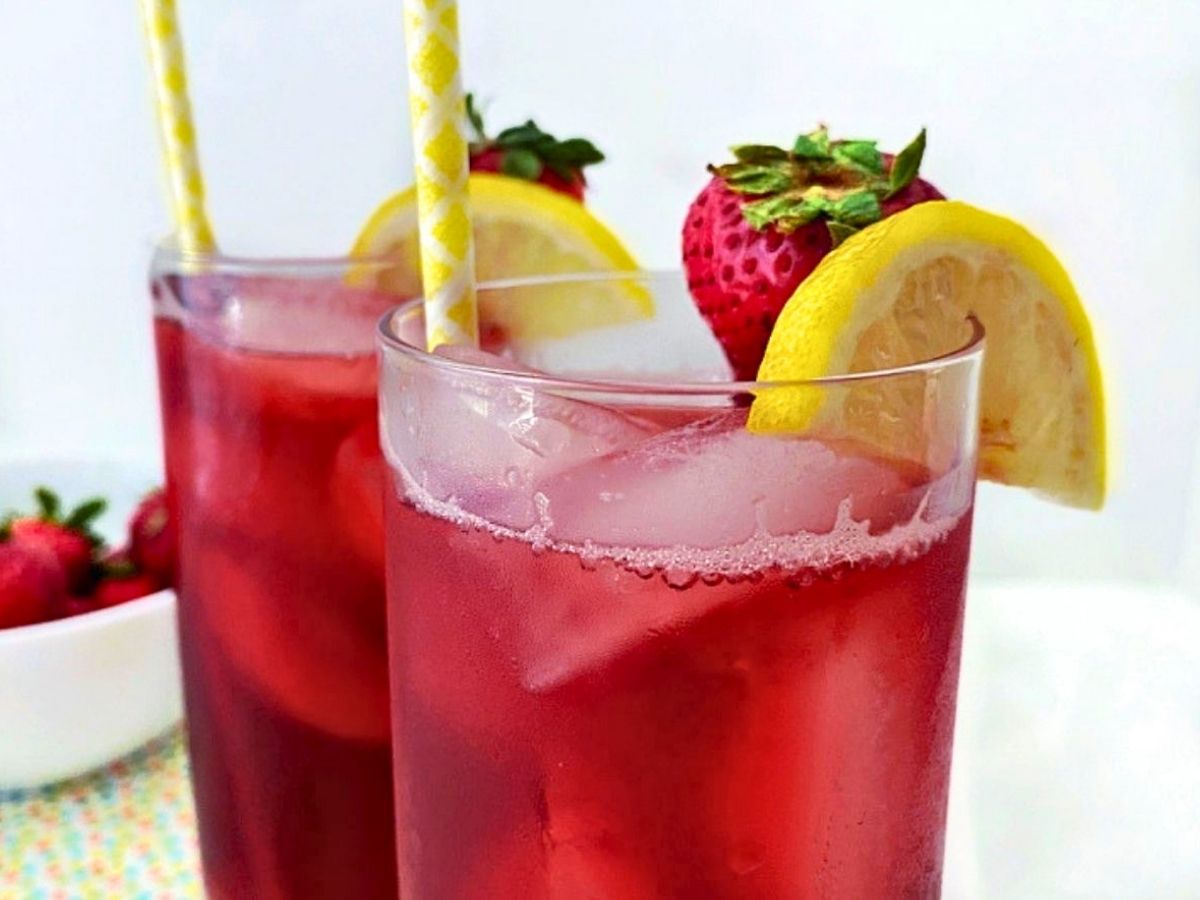 Two glasses of fruity iced tea with ice, paper straws, fresh strawberries, and lemon slices served on a colorful tabletop.