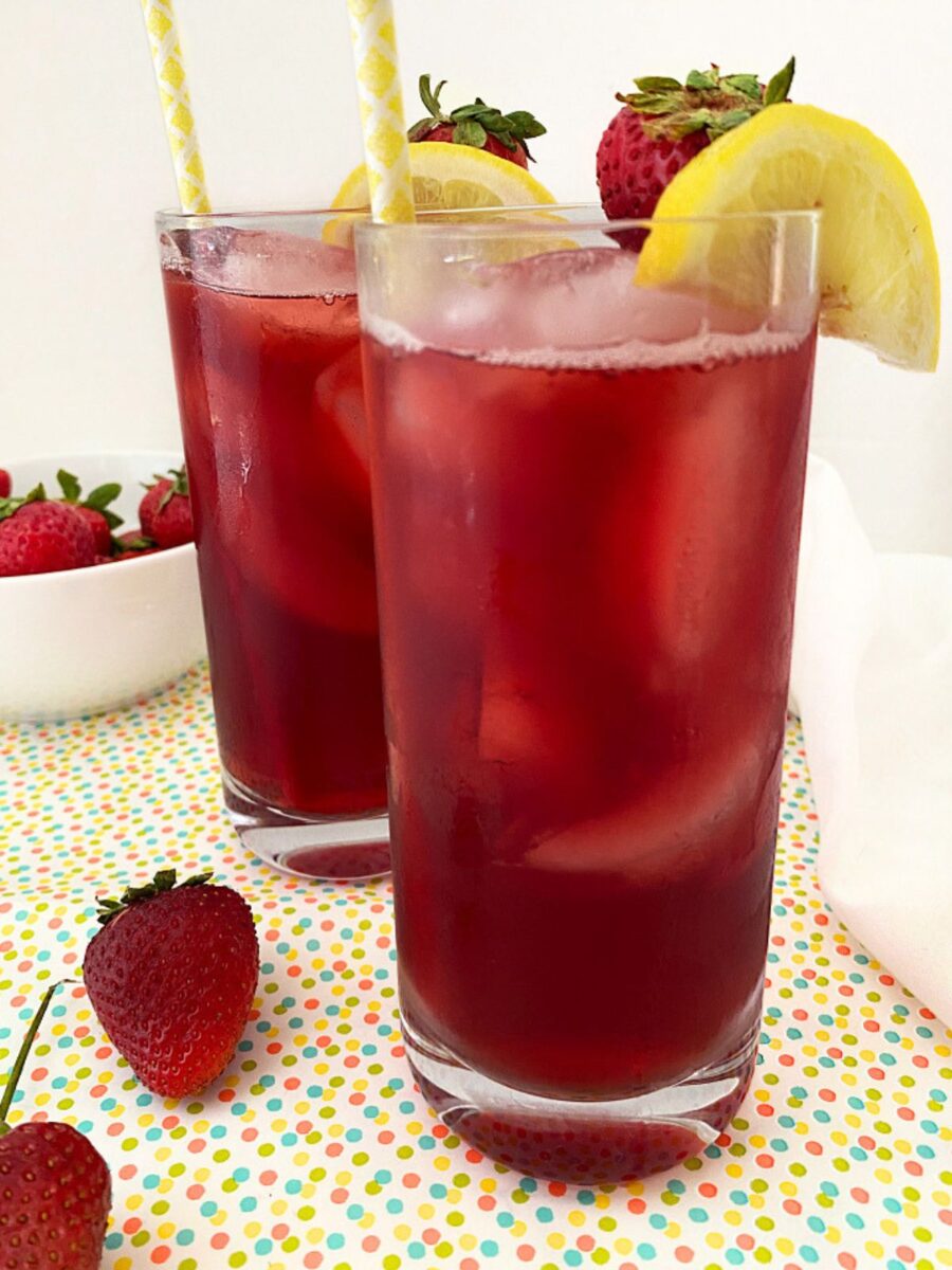 Two glasses of fruity iced tea with ice, paper straws, fresh strawberries, and lemon slices served on a colorful tabletop.