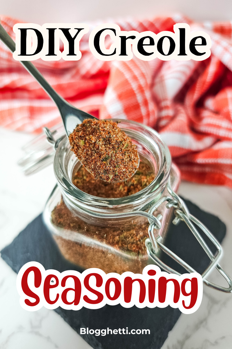 A close-up photo of a glass jar filled with homemade Creole seasoning. A spoonful of the spice blend is being lifted from the jar, showcasing its vibrant color and texture. The image is set against a rustic background with a red and white checkered cloth.