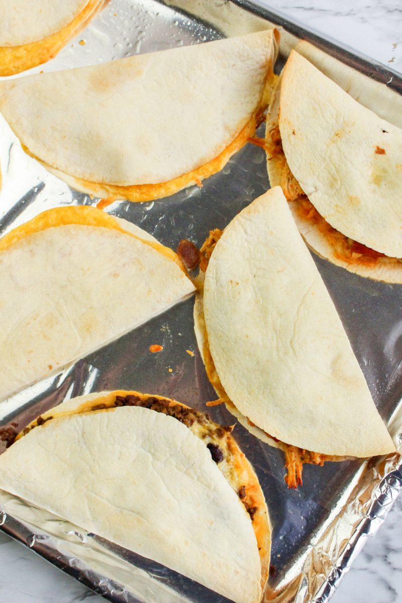 A baking sheet filled with folded quesadillas in various stages of browning. The quesadillas are filled with a mixture of cheese and other ingredients, visible through the open edges.