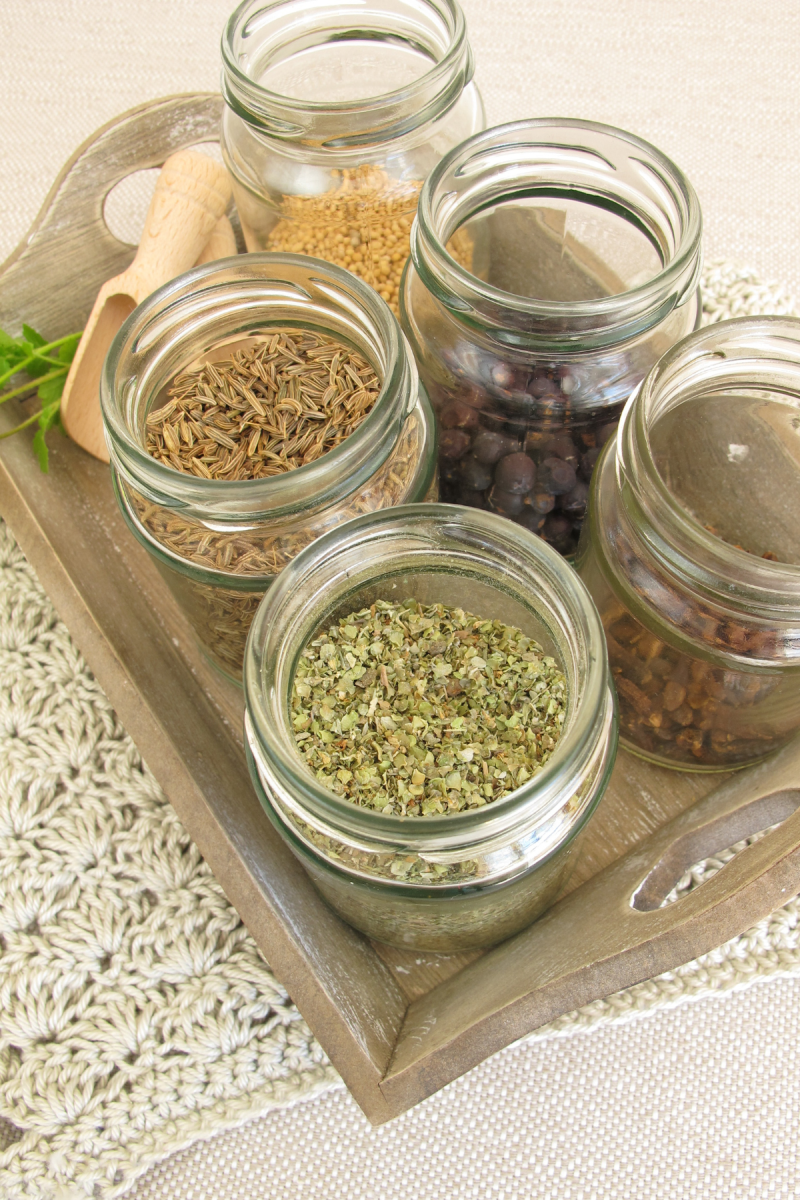 A wooden tray filled with glass jars of various spices. The jars contain cumin seeds, mustard seeds, juniper berries, dried oregano, and dried chili flakes. A small wooden scoop rests on the tray, and a sprig of parsley is in the background.