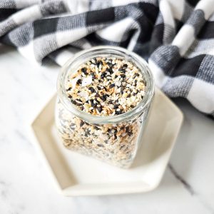 A jar filled with homemade everything bagel seasoning, made up of sesame seeds, poppy seeds, onion flakes, garlic powder, and other spices. The jar is sitting on a white plate with a black and white checkered cloth in the background.