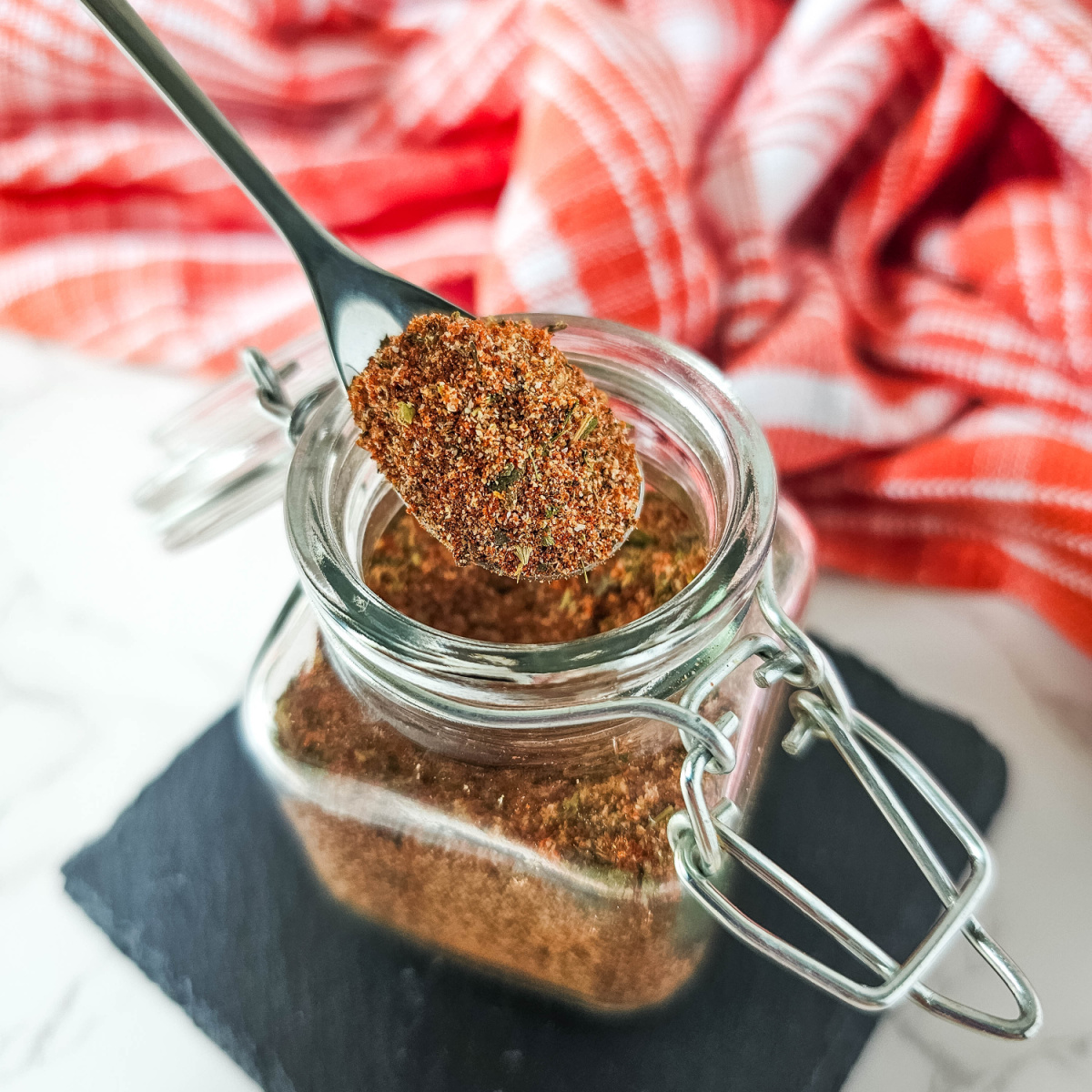 A close-up photo of a glass jar filled with homemade Creole seasoning. A spoonful of the spice blend is being lifted from the jar, showcasing its vibrant brown color and texture. The image is set against a rustic background with a red and white checkered cloth.