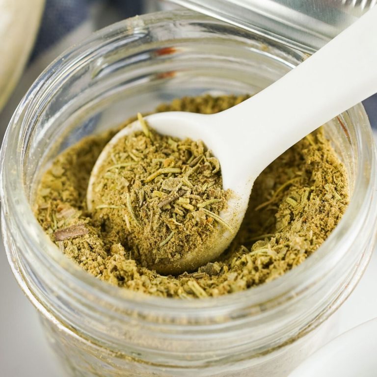 A close-up photo of a glass jar filled with homemade poultry seasoning, with a white spoon scooping some out. The seasoning is a brown mixture of herbs and spices.