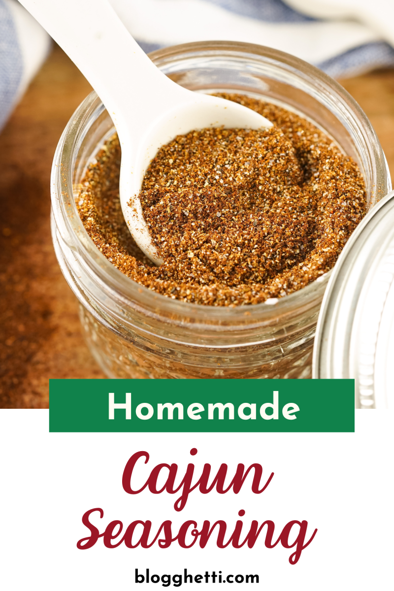 A jar of homemade Cajun seasoning with a white spoon scooping some out. The seasoning is a reddish-brown color.