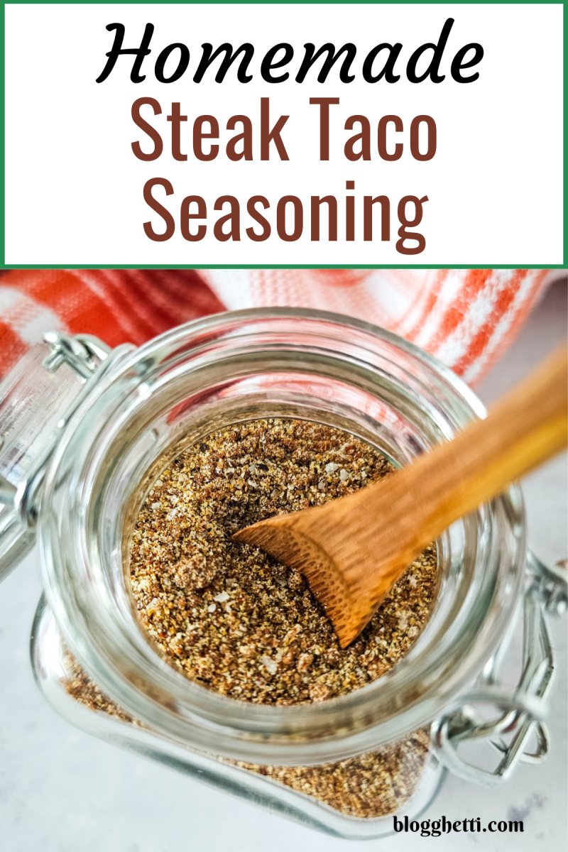 A close-up image of a glass jar filled with homemade steak taco seasoning. A small wooden spoon is scooping out some of the seasoning. The jar is sitting on a red and white checkered cloth.