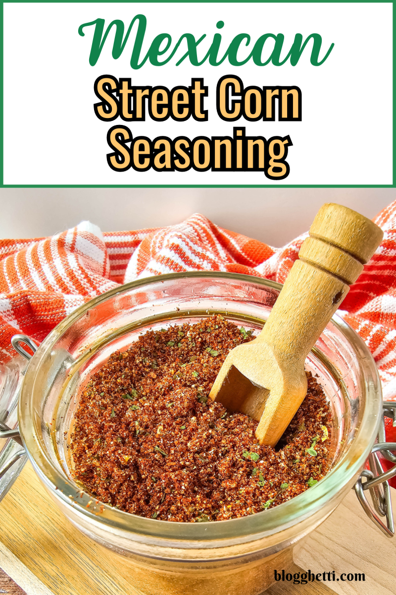 A jar of homemade Mexican street corn seasoning with a wooden scoop, sitting on a cutting board with a red and white striped cloth in the background.