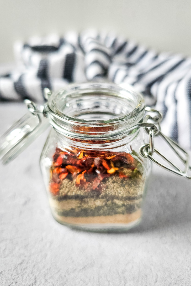 A glass jar with a metal clamp closure filled with a mixture of dried chili peppers, herbs, and spices. The jar is sitting on a gray surface with a striped cloth in the background.