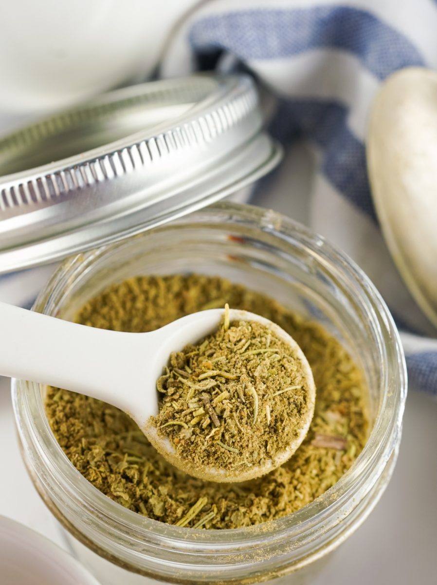 A close-up photo of a glass jar filled with homemade poultry seasoning, with a white spoon scooping some out. The seasoning is a brown mixture of herbs and spices.