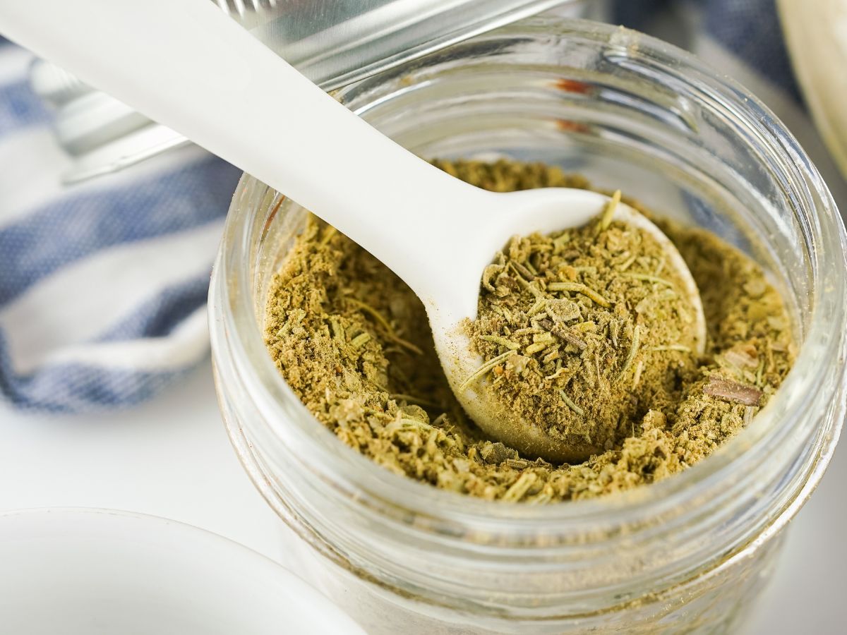 A close-up photo of a glass jar filled with homemade poultry seasoning, with a white spoon scooping some out. The seasoning is a brown mixture of herbs and spices.