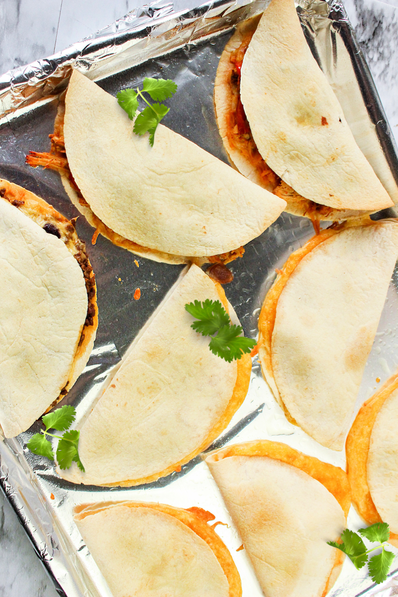 An overhead shot of a baking sheet filled with golden-brown, folded quesadillas. The quesadillas are filled with melted cheese and other ingredients, and garnished with fresh cilantro.