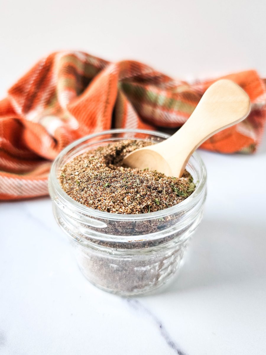 A close-up of a wooden spoon scooping up a flavorful seasoning blend from a glass bowl. The seasoning has a rich, brown color and appears to contain various herbs and spices.