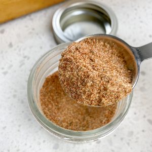 A jar of homemade Cajun seasoning with a white spoon scooping some out. The seasoning is a reddish-brown color.