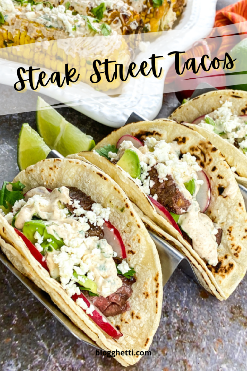 A plate of steak street tacos with grilled steak, avocado, cheese, radishes, and a creamy sauce on corn tortillas. There are lime wedges and a platter of grilled corn on the side.