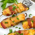 Four bacon-wrapped jalapeño poppers filled with melted cheese and topped with fresh parsley on a white rectangular plate. The image features a green header that reads "Bacon-Wrapped Jalapeño Poppers" and a circular "Easy Party Appetizer" badge.