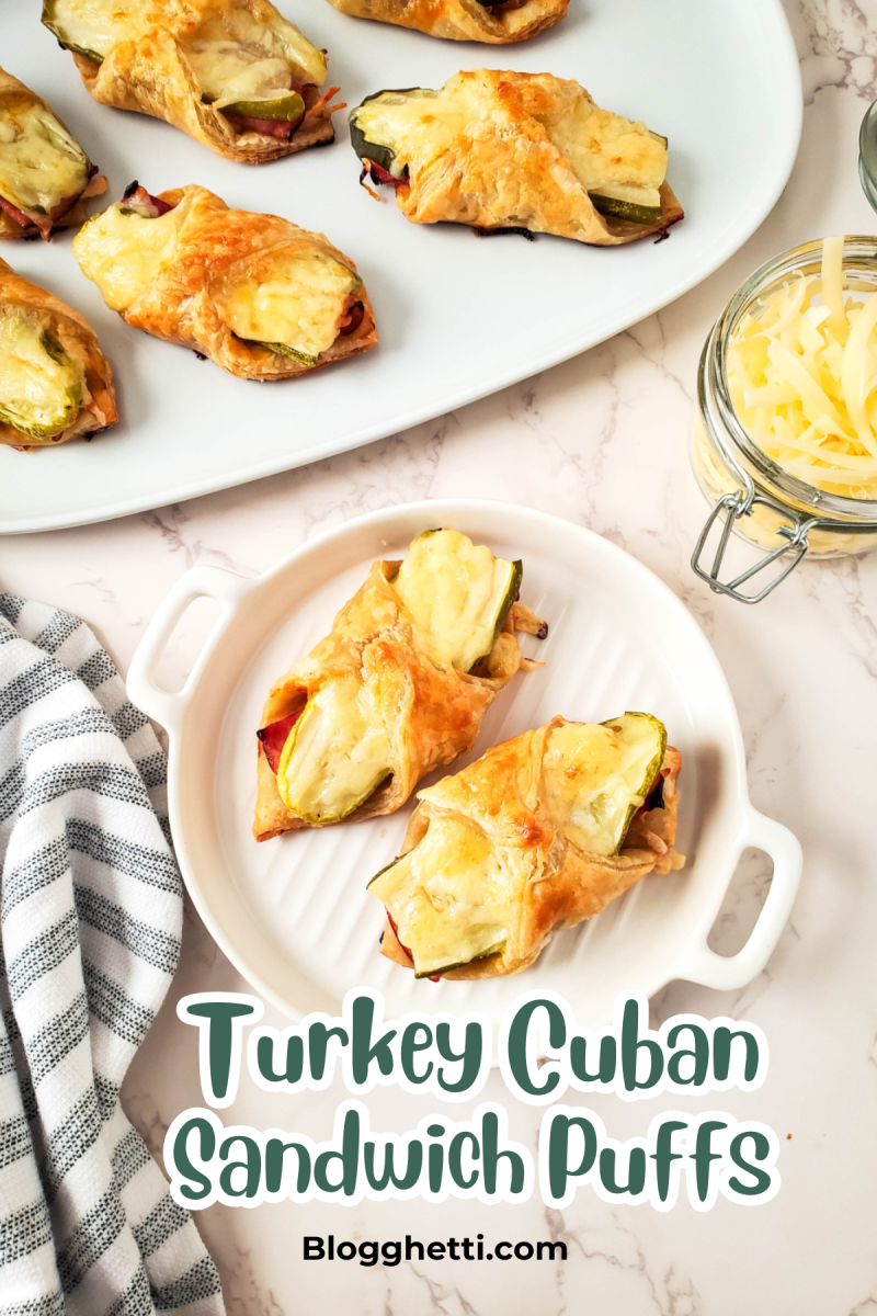 Overhead view of Turkey Cuban Sandwich Puffs served on a white plate and a white baking dish. These puffs are made with flaky pastry filled with turkey, Swiss cheese, and pickles, topped with melted cheese. A jar of shredded cheese and a striped kitchen towel are also visible in the setup. The text 'Turkey Cuban Sandwich Puffs' and 'Blogghetti.com' appear as an overlay on the image.