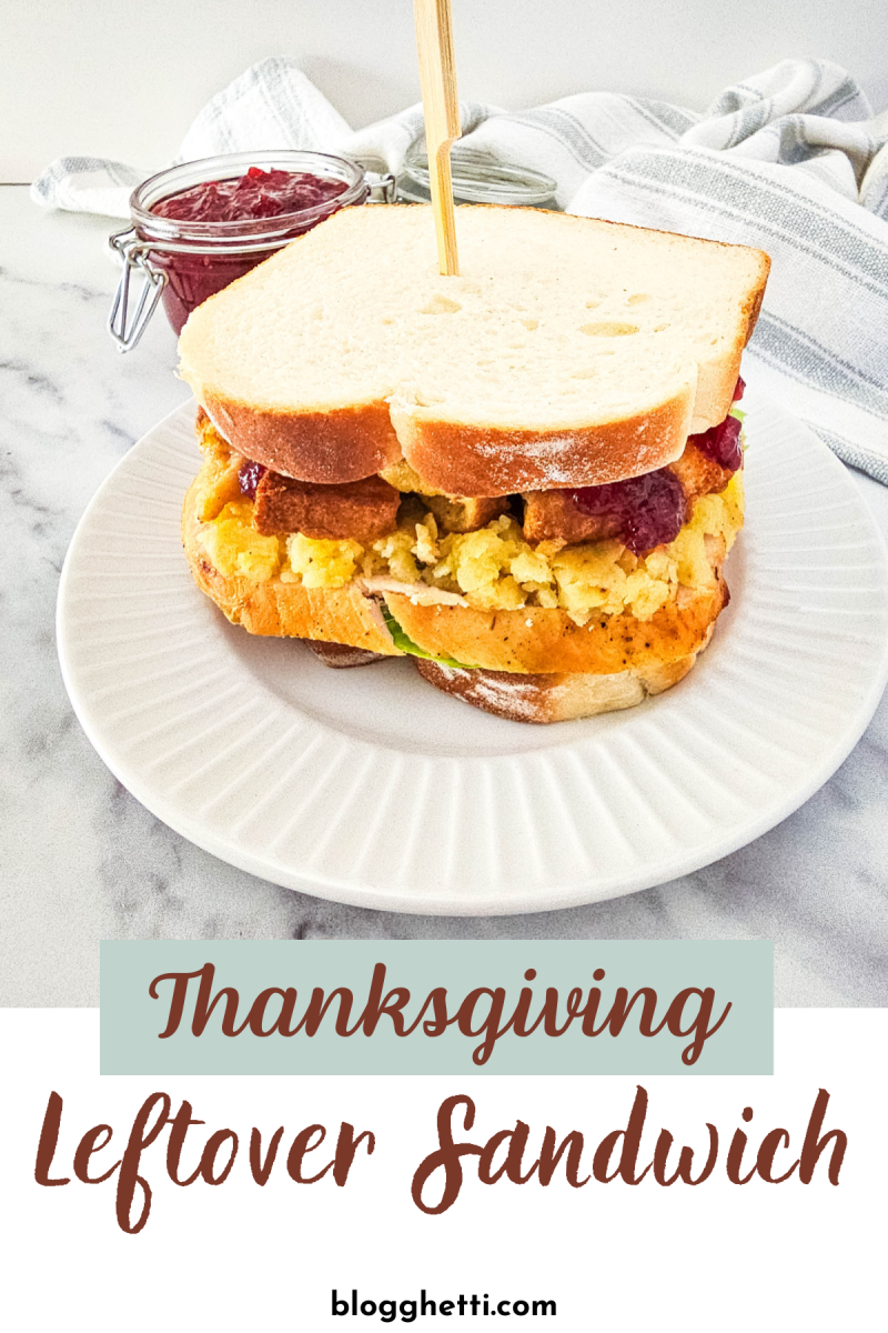 A delicious Thanksgiving leftover sandwich on a white plate. The sandwich is filled with turkey, mashed potatoes, stuffing, and cranberry sauce served on thick sliced bread. There is a jar of cranberry sauce on the side.