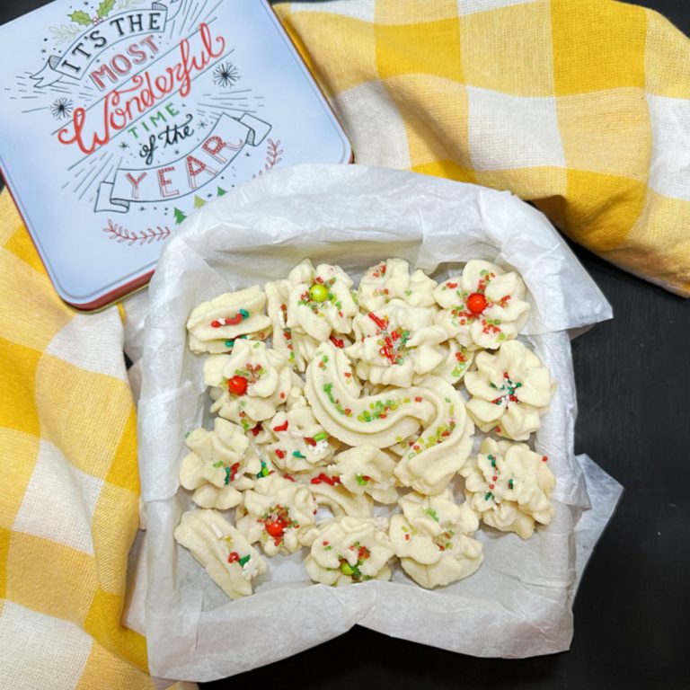 A festive display of Xianodianxin Eggless Chinese Butter Cookies decorated with colorful sprinkles, arranged in a parchment-lined tin, placed next to a holiday-themed container with the phrase "It's the Most Wonderful Time of the Year." A yellow checkered cloth adds a cheerful touch to the scene.